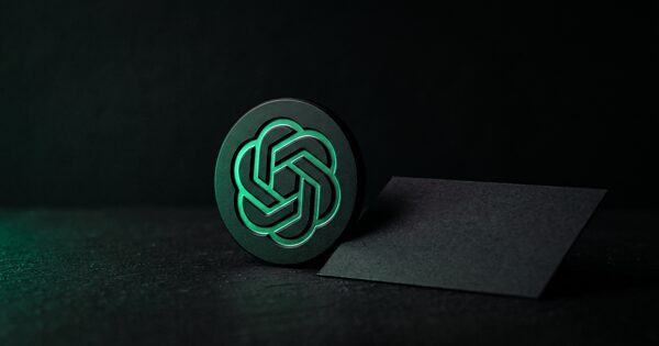Medalhão preto gravado com um nó de seis pétalas aceso a verde, com um pequeno cartão em branco encostado à base: os anúncios a entrar no ChatGPT